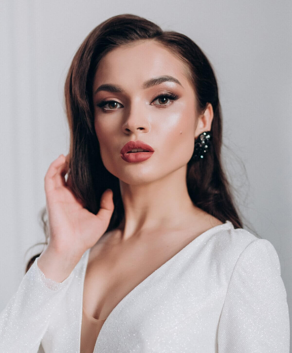 A lovely woman in a white dress poses with her hands on her face, radiating beauty and poise in a captivating manner.